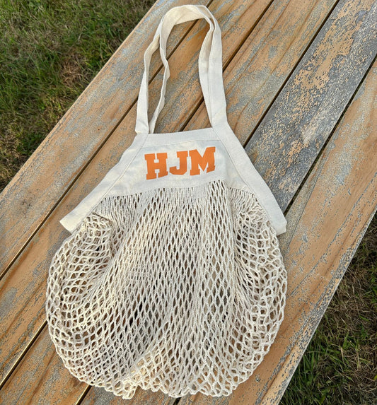 Mesh bag with 'HJM' branding on a wooden surface outdoors