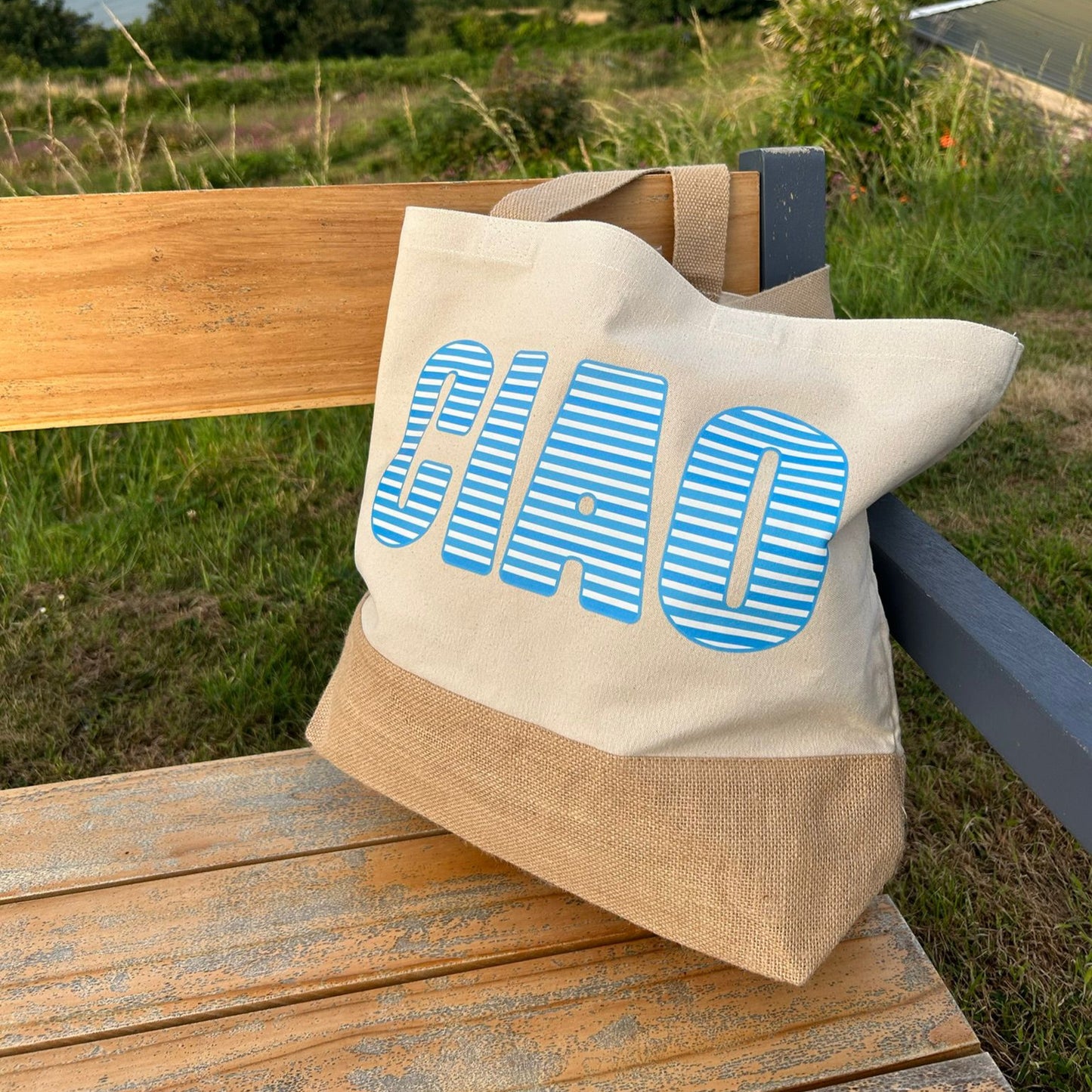 Tote bag with 'CIAO' printed on it, leaning against a wooden bench with a scenic background.